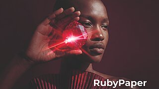 Nahaufnahme einer Person vor dunklem Hintergrund, die einen großen, facettierten Rubin vor das rechte Auge hält. Der rote Edelstein reflektiert ein helles Licht mit strahlenförmigen Lichteffekten. Die Person trägt ein gefaltetes Kleidungsstück in Burgunderrot